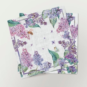 Può includere: Un set di tre tovaglioli di lino con un design floreale che presenta fiori di lillà viola e bianco, foglie verdi, un'ape e una farfalla. I tovaglioli hanno uno sfondo bianco con una sottile trama di lino.