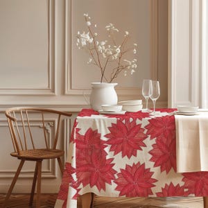 May include: A rectangular tablecloth featuring a repeating pattern of large, vibrant red poinsettia flowers on a cream-coloured background. The fabric drapes over a table set with a white pitcher, white bowls, and two clear glass wine stems.