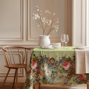 Könnte beinhalten: Ein Esstisch mit einer floralen Tischdecke mit Rosen und anderen Blumen in Rosa-, Lila- und Weißtönen. Der Tisch ist mit weißem Geschirr, Weingläsern und einer weißen Vase mit weißen Blumen gedeckt.