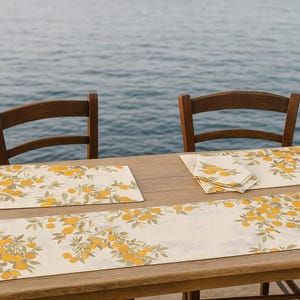 May include: A wooden table set with a cream-coloured table runner, placemats, and napkins. The linens feature a repeating pattern of yellow citrus fruit and green leaves. Two wooden chairs are visible in the background, with the ocean in the distance.