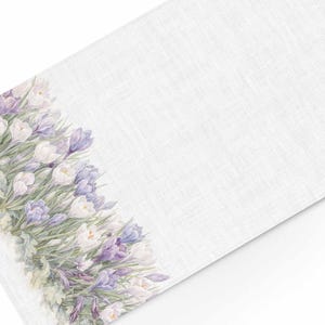 Peut inclure: Une nappe rectangulaire blanche avec un motif floral à l'aquarelle. Le motif présente des grappes de fleurs de crocus violettes et blanches avec un feuillage vert le long d'un bord. Le fond est un tissu blanc texturé.