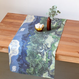 Puede incluir: Un camino de mesa azul y verde con un patrón de remolino, que recuerda a una pintura famosa. El camino de mesa está sobre una mesa de madera con un pequeño jarrón de vegetación y un tazón de malvaviscos blancos.