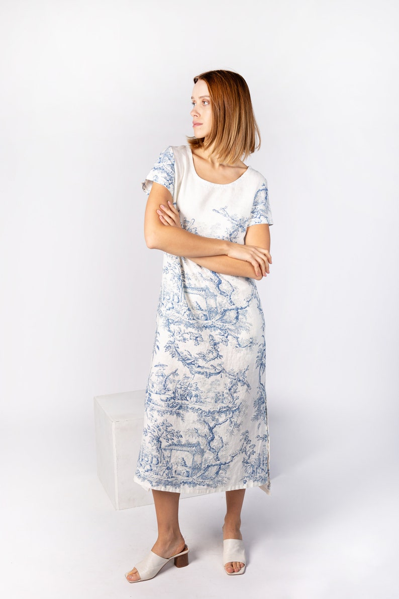 May include: A white linen dress with a blue and white floral print. The dress has a round neckline and short sleeves. The model is wearing white sandals.