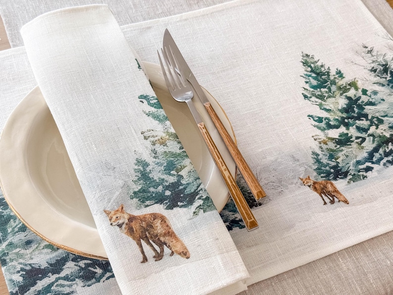 May include: A white linen placemat with a winter forest scene featuring green pine trees and a red fox. The placemat is set with a white plate and a silver fork and knife with wooden handles.