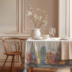 Può includere: Una tovaglia bianca con un designo di città colorato che presenta case, un ponte e un fiume. La tovaglia è posta su un tavolo di legno con un vaso bianco di fiori bianchi, due bicchieri di vino e ciotole.