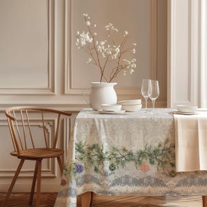 Op de afbeelding: Een rechthoekig tafelkleed met een geprint patroon van groene dennentakken en kleurrijke ronde ornamenten op een lichtgrijze, kantachtige achtergrond. De tafel is gedekt met een witte kan, witte kommen en twee heldere glazen flutes.