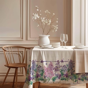 May include: A white tablecloth with a floral pattern of purple and green lilac flowers. The tablecloth is set on a wooden table with a white vase of white flowers, two wine glasses, and white bowls.