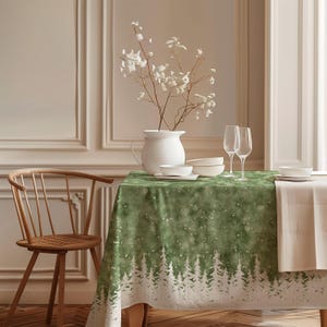 Pode incluir: Uma mesa de jantar com uma toalha de mesa verde com um desenho de floresta branca. A mesa está posta com louça branca, taças de vinho e um vaso branco com flores brancas. Uma cadeira de madeira está à esquerda.