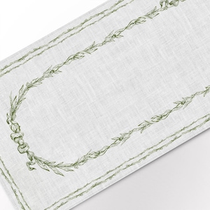 Op de afbeelding: Een witte linnen tafelrunner met een groene, bladerige randontwerp. De rand heeft een herhalend patroon van bladeren en bessen.