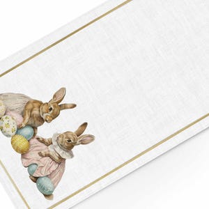 Può includere: Runner da tavolo pasquale bianco con bordo dorato. Il runner presenta due coniglietti in abito, circondati da uova di Pasqua dai colori pastello. I coniglietti sono marroni e beige, e le uova sono decorate con motivi floreali.