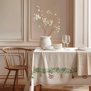 May include: A white linen tablecloth with a green garland border and Christmas ornaments. The tablecloth is set on a wooden table with a white vase of dried flowers, two wine glasses, and white bowls.