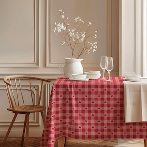 May include: A red and pink checkered tablecloth with heart patterns covers a table. A white vase with white flowers sits on the table, along with white bowls, plates, and wine glasses. A wooden chair is on the left.