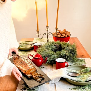 May include: A table setting with a smoked fish on a slate platter, a red teapot, two red mugs, a green bowl, a silver candle holder with three yellow candles, a bowl of walnuts, and a pine wreath. The table is covered with a linen tablecloth with a green pine tree pattern.