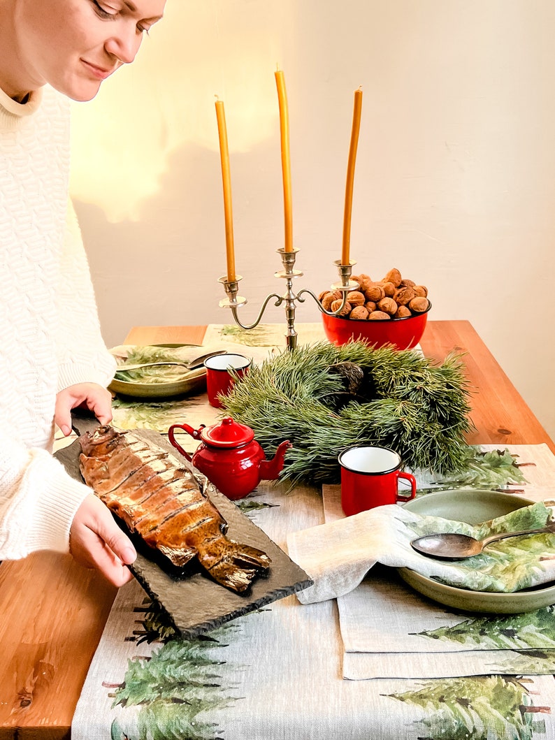 May include: A table setting with a smoked fish on a slate platter, a red teapot, two red mugs, a green bowl, a silver candle holder with three yellow candles, a bowl of walnuts, and a pine wreath. The table is covered with a linen tablecloth with a green pine tree pattern.