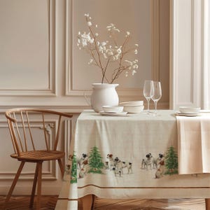 Puede incluir: Un mantel rectangular de color beige con un patrón repetitivo de perros pequeños y pinos verdes a lo largo del borde inferior. La tela tiene una apariencia texturizada y cubre una mesa con una jarra blanca, cuencos y dos copas.