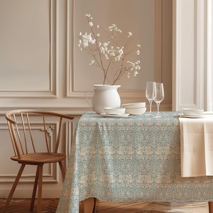 Pode incluir: Uma toalha de mesa retangular com um padrão botânico repetitivo em azul-petróleo e creme, com motivos intrincados de flores e vinhas. O tecido cobre uma mesa posta com um jarro de cerâmica branca, taças brancas e duas flutes de vidro.