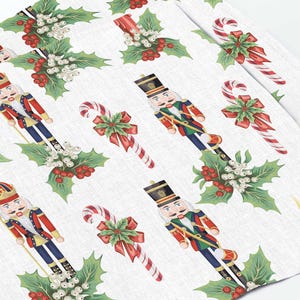 Könnte beinhalten: Weißer Hintergrund mit einem sich wiederholenden Muster aus Nussknackern, Zuckerstangen und Stechpalmen. Die Nussknacker tragen rote und blaue Uniformen und halten eine Zuckerstange. Die Zuckerstangen sind rot und weiß und die Stechpalme ist grün mit roten Beeren.