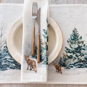 May include: A white linen placemat with a winter forest scene featuring pine trees and foxes. The placemat has a white napkin with a fox design and a fork and knife on top.