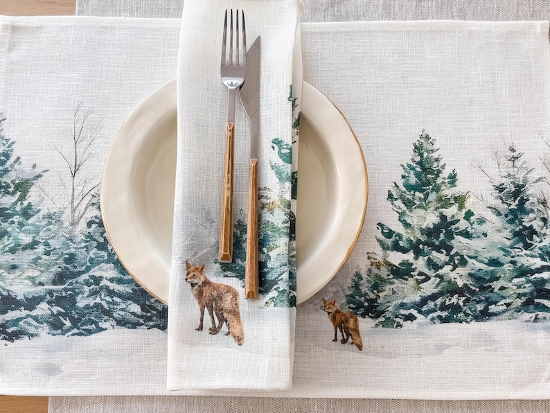 May include: A white linen placemat with a winter forest scene featuring pine trees and foxes. The placemat has a white napkin with a fox design and a fork and knife on top.