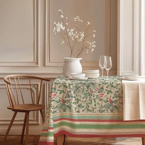 May include: A rectangular tablecloth featuring a botanical print of green holly leaves, pine needles, and red berries on a cream background, finished with horizontal stripes in red, green, and cream along the bottom edge of the fabric.