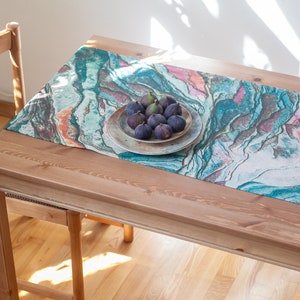 May include: A teal, blue, and pink marble-patterned table runner with a plate of figs on top. The runner is on a wooden table.