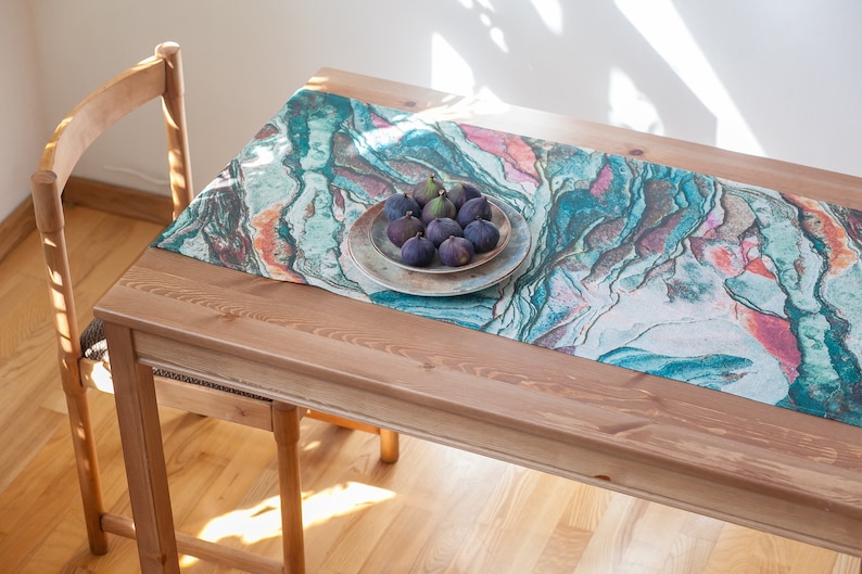 May include: A teal, blue, and pink marble-patterned table runner with a plate of figs on top. The runner is on a wooden table.