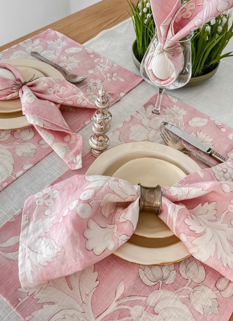 May include: Pink floral linen napkins with white embroidery, folded and placed on a pink floral linen placemat with a silver napkin ring. The placemat is on a light grey linen tablecloth.