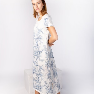 May include: A white linen dress with a blue floral print. The dress has a round neckline, short sleeves, and a side slit. The model is wearing white sandals.