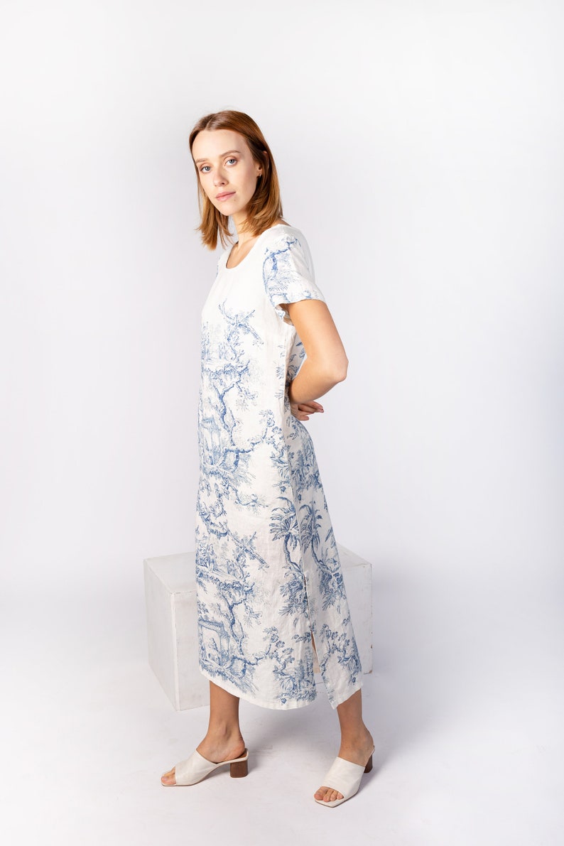May include: A white linen dress with a blue floral print. The dress has a round neckline, short sleeves, and a side slit. The model is wearing white sandals.