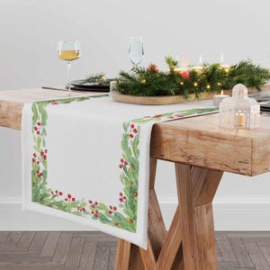 Può includere: Tovaglia da tavolo bianca con un bordo di agrifoglio verde e rosso. La tovaglia è su un tavolo di legno con un centrotavola di verde e candele.