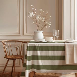 Può includere: Un tavolo da pranzo apparecchiato con una tovaglia a righe verdi e bianche, piatti bianchi e bicchieri da vino trasparenti. Un vaso bianco contiene fiori bianchi. Una sedia di legno è a sinistra.