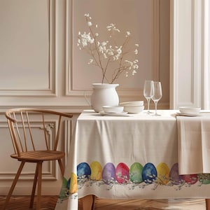 Peut inclure: Une nappe en lin blanc avec une bordure d'œufs de Pâques colorés. La nappe est posée sur une table en bois avec un vase blanc de fleurs blanches, deux verres à vin et des bols blancs.