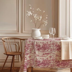 Pode incluir: Uma toalha de mesa retangular com um padrão repetitivo de toile de jouy vermelho escuro sobre um fundo creme. A mesa está posta com um jarro de cerâmica branca contendo flores brancas, duas taças de vidro transparente e várias tigelas.