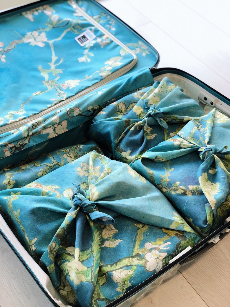 K&ouml;nnte beinhalten: Ein blauer Koffer mit einem floralen Muster als Innenfutter. Der Koffer ist ge&ouml;ffnet und enth&auml;lt vier Gegenst&auml;nde, die in blaues Stoff mit einem floralen Muster gewickelt sind. Der Stoff ist mit Knoten gebunden.