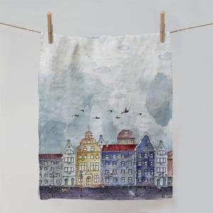 May include: A watercolour illustration of a city street with colourful buildings and birds flying overhead. The buildings are painted in shades of yellow, blue, and red, and the sky is a soft blue with white clouds. The illustration is printed on a linen tea towel.