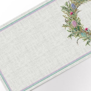May include: A rectangular linen table runner with an Easter wreath design. The runner has a light grey background with green and purple stripes along the edges. The wreath features green branches, colourful eggs, and berries.