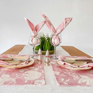 May include: Two pink floral placemats with white floral designs, each with a white egg wrapped in a pink napkin folded into bunny ears, a plate, and a silver knife. The placemats are on a white linen tablecloth with a wooden table in the background.