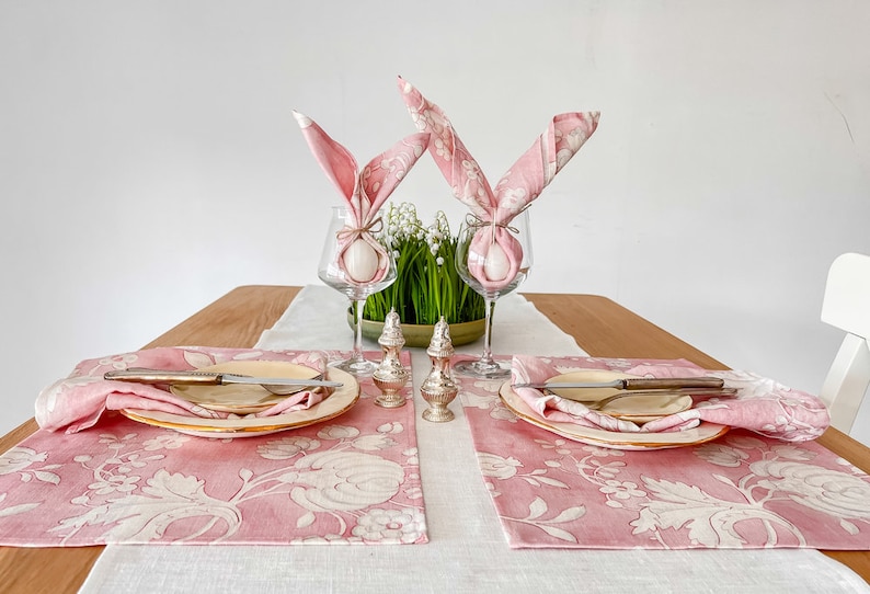 May include: Two pink floral placemats with white floral designs, each with a white egg wrapped in a pink napkin folded into bunny ears, a plate, and a silver knife. The placemats are on a white linen tablecloth with a wooden table in the background.