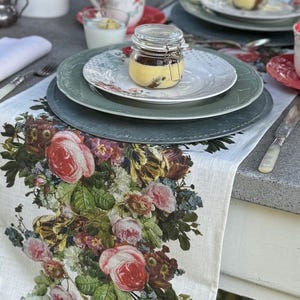 Könnte beinhalten: Ein weißer Tischläufer aus Leinen mit einem Blumendruck mit rosa, roten und gelben Rosen. Der Läufer liegt auf einem Tisch mit zwei Gedecken, jedes mit einem grünen Teller, einem weißen Teller mit Blumendekor und einem Glasgefäß gefüllt mit einem gelben Dessert.