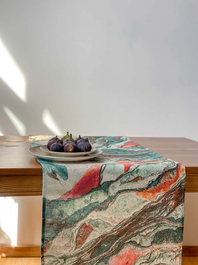 May include: A green, orange, and red abstract marble patterned table runner on a wooden table with a white plate of figs on top.