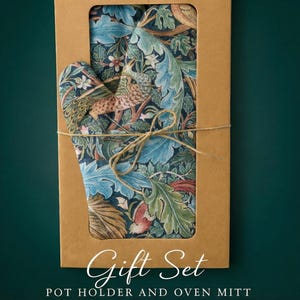 Puede incluir: Un juego de regalo con un salvamanteles y un guante de horno. Los artículos se muestran en una caja de cartón marrón con una ventana transparente, atados con hilo. La tela tiene un estampado floral y de pájaros azul, verde y marrón. Texto: "Gift Set POT HOLDER AND OVEN MITT."