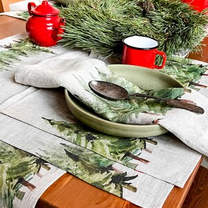 May include: A set of two linen placemats with a green watercolor pine tree design. The placemats are set on a wooden table with a green wreath, a red teapot, and a red enamel mug.