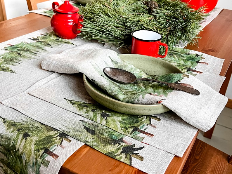 May include: A set of two linen placemats with a green watercolor pine tree design. The placemats are set on a wooden table with a green wreath, a red teapot, and a red enamel mug.