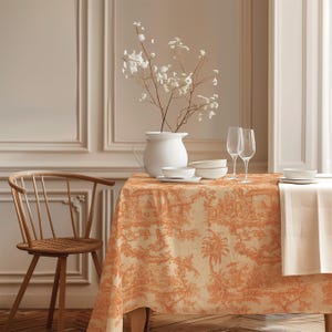 May include: A rectangular tablecloth featuring an orange and cream toile de jouy pattern with pastoral scenes. On the table sits a white ceramic pitcher containing white flowers, two clear glass flutes, and stacked white ceramic bowls.