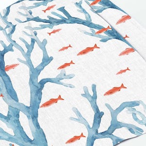 Pode incluir: Um tecido branco com um padrão de aquarela de coral azul e peixes laranja. O tecido tem uma textura de linho sutil.