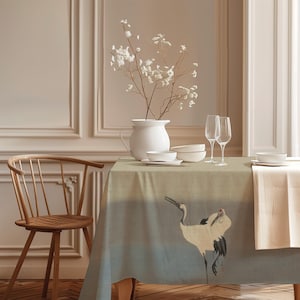 Könnte beinhalten: Ein Esstisch mit einer salbeigrünen Tischdecke mit einer Kranichillustration. Der Tisch ist mit weißem Geschirr, Weingläsern und einer weißen Vase mit weißen Blumen gedeckt. Links steht ein Holzstuhl.