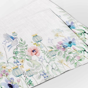 May include: A white linen napkin with a watercolor floral design. The design features blue, pink, and green flowers, leaves, and a small blue bird.