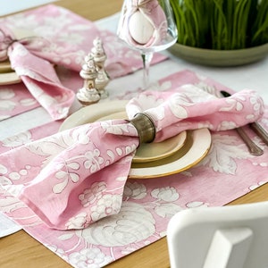 May include: Pink floral placemats with white designs, a pink napkin with a silver napkin ring, and a white egg wrapped in a pink napkin.