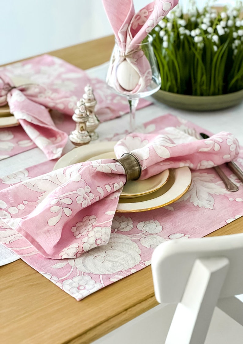 May include: Pink floral placemats with white designs, a pink napkin with a silver napkin ring, and a white egg wrapped in a pink napkin.
