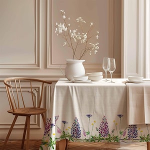 May include: A white linen tablecloth with a floral border featuring purple and green wildflowers. The tablecloth is set on a wooden table with two white bowls, two wine glasses, and a white pitcher with white flowers.